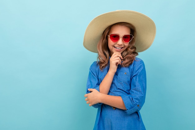 Menina bonita modelo com cabelos castanhos ondulados em vestido, chapéu grande, sorrisos de óculos de sol vermelhos isolados em azul