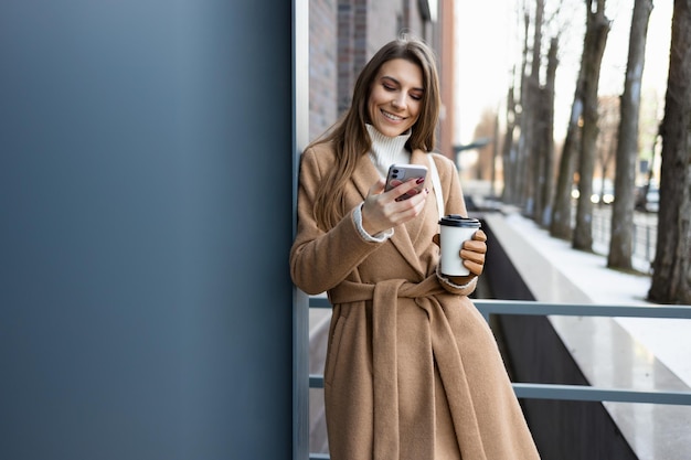 Menina bonita morena com casaco marrom com xícara de café usando smartphone ao ar livre