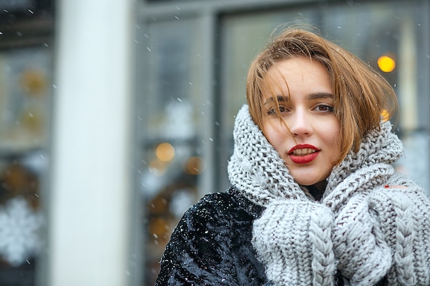 Menina bonita morena com lábios vermelhos usa casaco e lenço andando na cidade durante a queda de neve. Espaço vazio