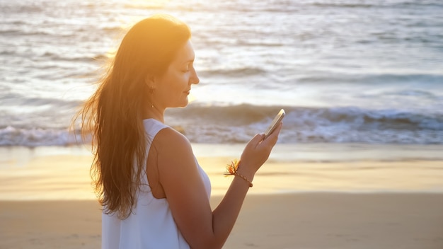 Menina bonita morena digitando em um smartphone na praia do oceano ao pôr do sol