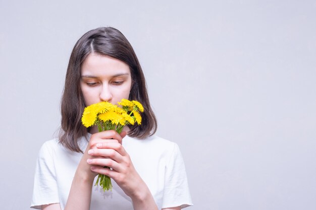 Menina bonita morena segurando flores silvestres amarelas de leão e inalando seu aroma sobre um fundo claro, copie o espaço. Flores silvestres de primavera. Amor, romance, conceito de casamento