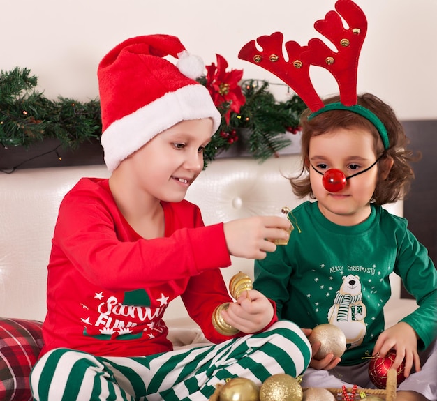 Menina bonita na máscara de veado engraçado e o menino no chapéu de Papai Noel com pijama de Natal tirar os brinquedos da caixa e se preparar para o feriado.