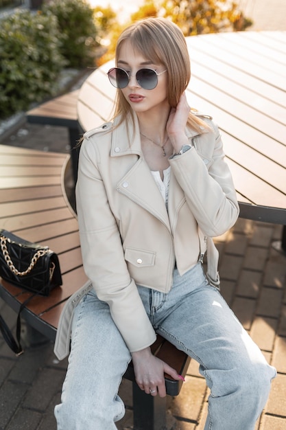 Menina bonita na moda jovem hippie com óculos de sol elegantes em uma roupa de moda rock com uma jaqueta de couro e jeans azul com bolsa sentado em um banco de madeira ao pôr do sol Roupas urbanas femininas