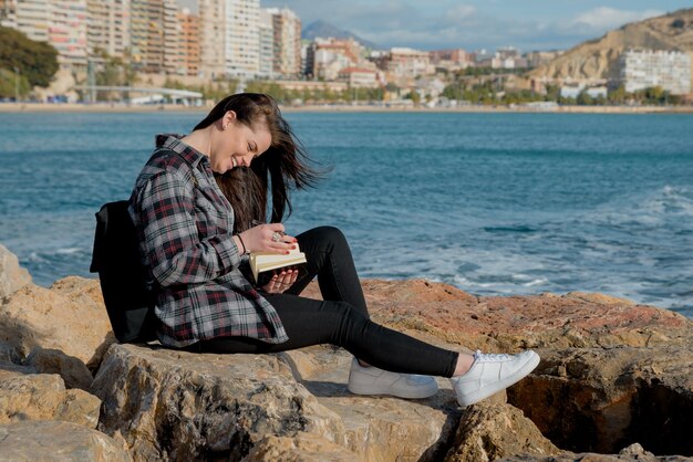 Menina bonita nas rochas da praia ao ar livre escreve histórias e pensamentos para seu livro em seu caderno