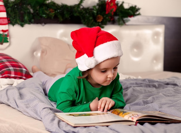 Menina bonita no chapéu de Papai Noel e pijama de Natal, lendo um livro.