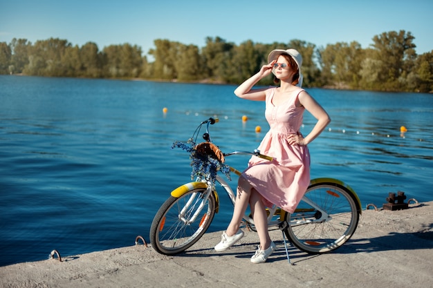 Menina bonita no chapéu e óculos de sol em uma bicicleta na margem do rio. A menina olha para longe