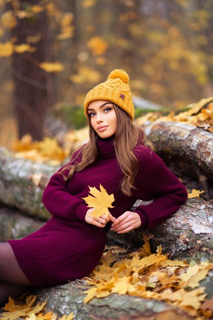 Menina bonita no confortável vestido de malha cor de vinho e chapéu sentado na natureza com fundo de outono.