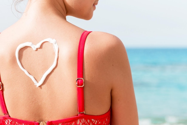 Menina bonita no maiô vermelho tem protetor solar em forma de coração na parte de trás na praia