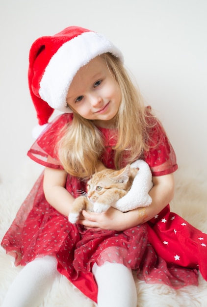 Menina bonita no vestido de festa vermelho de chapéu de Papai Noel com gatinho de gengibre esperando o Natal