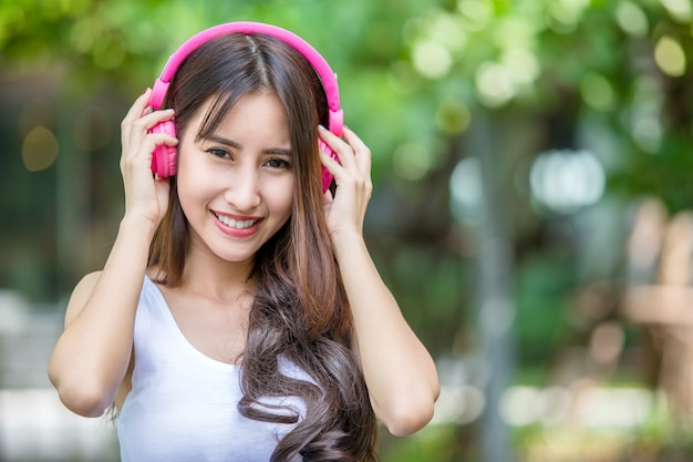 Menina bonita nova feliz que escuta a música com seus auscultadores cor-de-rosa e que dança no parque urbano