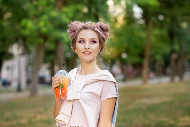 Menina bonita nova que bebe o suco fresco dos copos afastados plásticos do alimento após uma caminhada fora. estilo de vida saudável. sorrindo magro loira com cabelo rosa.