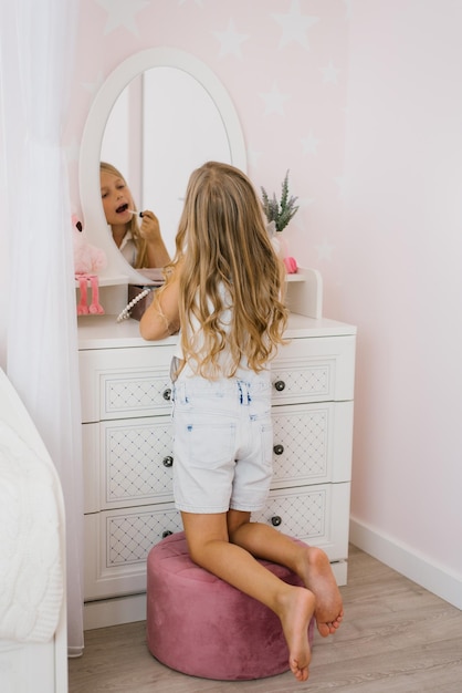 Menina bonita pinta os lábios com glitter ou bálsamo na frente de um espelho em seu quarto
