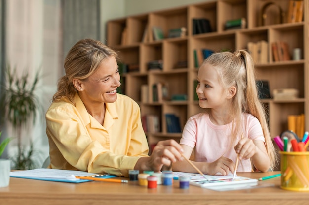 Menina bonita pintando com especialista em crianças sentada à mesa com professora e segurando espaço de cópia de pincel