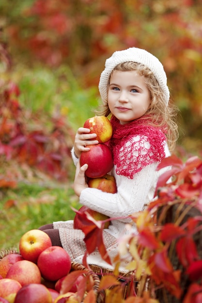 Menina bonita que guarda maçãs no jardim do outono. . Menina brincando no pomar de árvores de maçã. Criança comendo frutas na colheita de outono. Diversão ao ar livre para crianças. Nutrição saudável