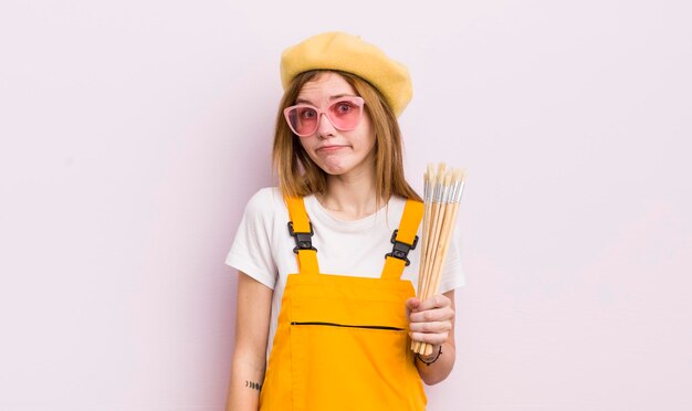 Menina bonita ruiva encolhendo os ombros sentindo-se confuso e incerto conceito de estudante de arte