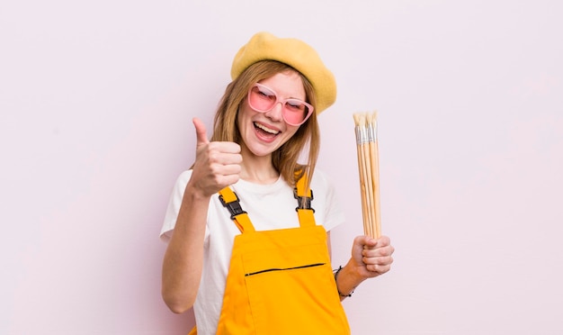 Menina bonita ruiva se sentindo orgulhosa sorrindo positivamente com polegares para cima conceito de estudante de arte