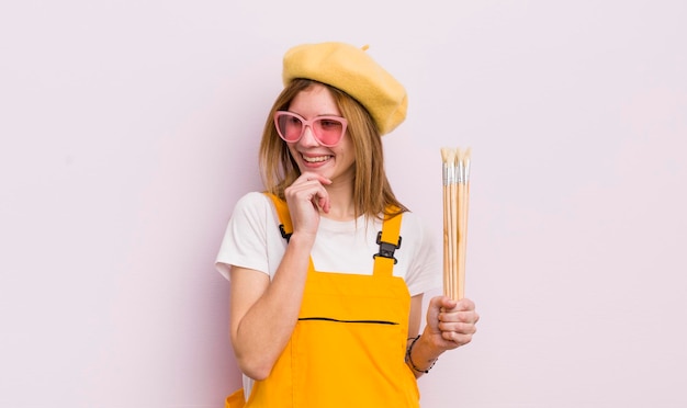 Menina bonita ruiva sorrindo com uma expressão feliz e confiante com a mão no conceito de estudante de arte do queixo