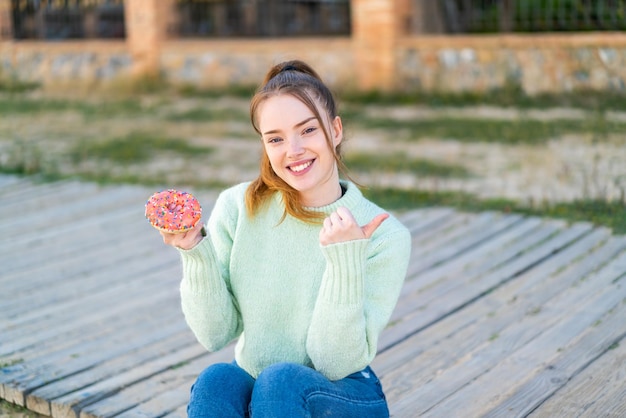 Menina bonita segurando uma rosquinha ao ar livre apontando para o lado para apresentar um produto