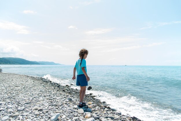 Menina bonita senta-se em uma praia de seixos e brinca com seixos