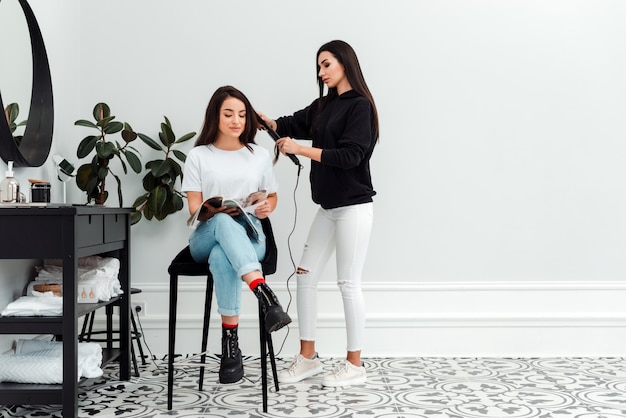 Menina bonita sentada na cadeira de cabeleireiro, lendo revista, seu mestre fazendo penteado