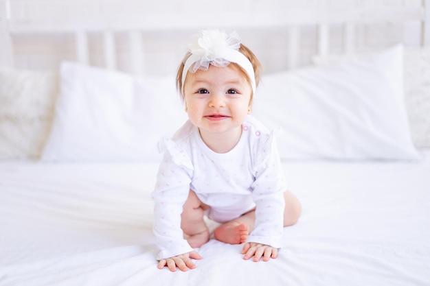 Menina bonita sentada na cama com roupas brancas e com um laço na cabeça bebezinho engraçado em uma cama de algodão em casa e sorrindo o conceito de produtos de bebê