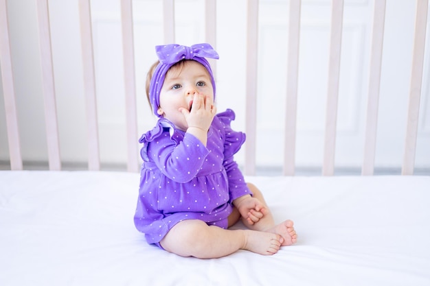 Menina bonita sentada na cama com roupas lilás e com um laço na cabeça e sorrindo bebê engraçado em uma cama de algodão em casa jogando o conceito de produtos infantis