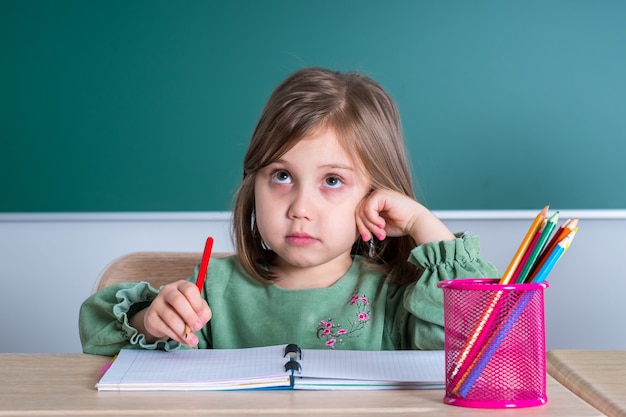 Menina bonita sentada na sala de aula na mesa cinza e desenha.