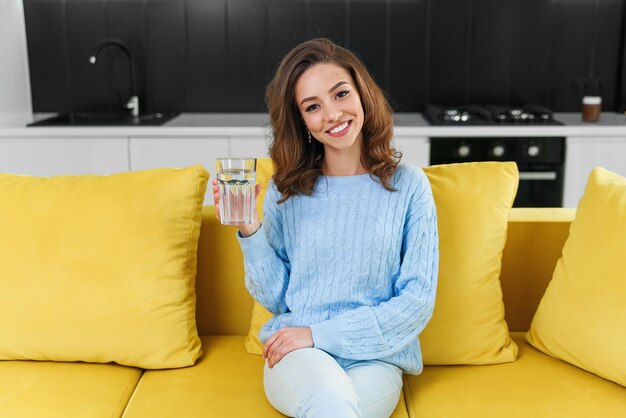 Menina bonita sentada no confortável sofá amarelo e bebe um copo de água pura no