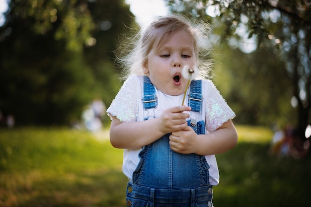 Menina bonita soprando sementes de dente de leão no pôr do sol no parque de verão Imagem com foco seletivo