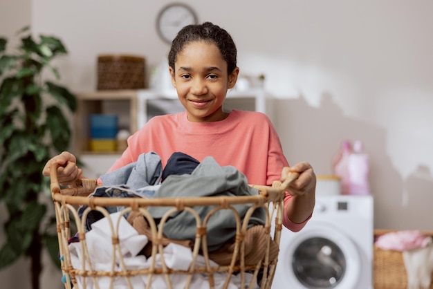 Menina bonita sorridente fica no meio da lavanderia do banheiro segurando uma grande cesta de vime cheia de roupas coloridas nas mãos filha ajuda a mãe com as tarefas domésticas