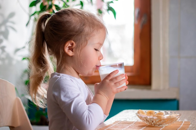 Menina bonita tomando café da manhã com pedaços de banana e copo de leite pela manhã