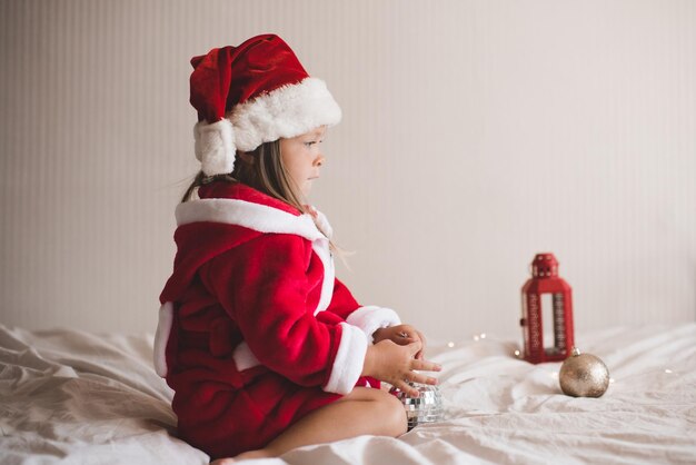 Menina bonita usa chapéu de papai noel vermelho e roupão de banho sentado na cama com decoração de natal