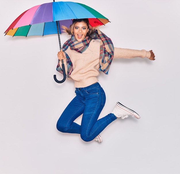 Menina bonita vestindo roupas de inverno e cachecol sorrindo feliz pulando com sorriso no rosto segurando guarda-chuva colorido sobre fundo branco isolado