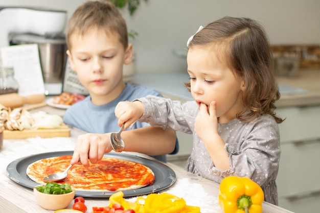 Menina bonitinha 2-4 em vestido cinza e menino 7-10 em t-shirt cozinhando pizza juntos na cozinha. irmão e irmã cozinhando