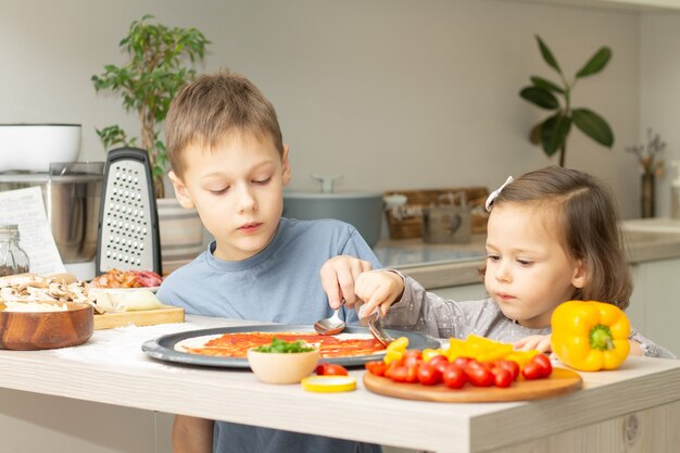 Menina bonitinha 2-4 em vestido cinza e menino 7-10 em t-shirt cozinhando pizza juntos na cozinha. Irmão e irmã cozinhando
