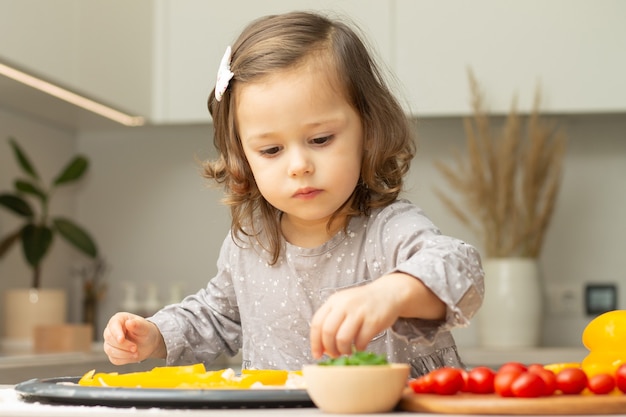 Menina bonitinha 2-4 no vestido cinza, cozinhar pizza na cozinha. kid arruma ingredientes em uma base de pizza