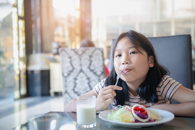 Menina bonitinha asiática comendo tomate fresco e salada de manhã no hotel. Saudável e relaxe no conceito de férias