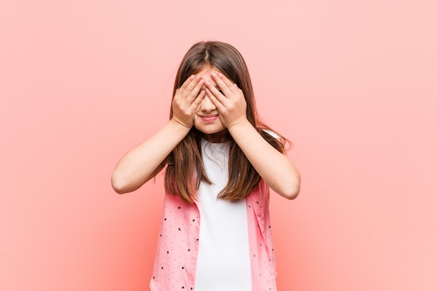 Menina bonitinha cobre os olhos com as mãos, sorri amplamente esperando por uma surpresa.