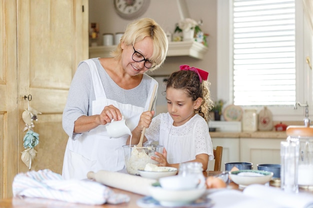 Menina bonitinha com a avó cozinhando juntos na mesa da cozinha. Visualização horizontal.