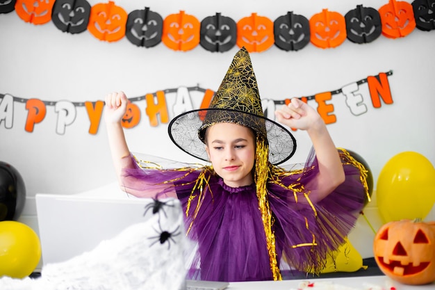 Menina bonitinha com chapéu de bruxa sentada atrás da mesa no quarto decorado com tema de Halloween, Covid19
