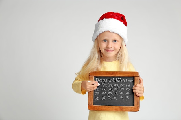 Menina bonitinha com chapéu de Papai Noel com quadro-negro contando os dias até o Natal, sobre fundo claro