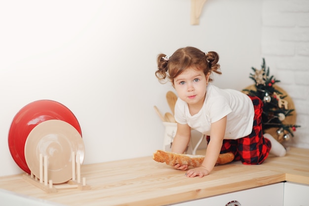 Menina bonitinha com chapéu de papai noel, preparando biscoitos na cozinha em casa. senta-se na mesa da cozinha e ajuda a mãe a preparar um jantar festivo de natal