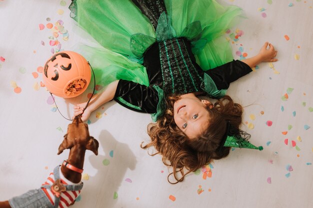 Menina bonitinha com fantasia de Halloween verde deitada no chão coberta de confetes coloridos