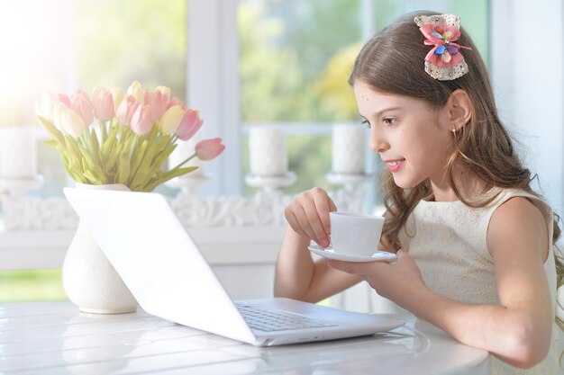 Menina bonitinha com laptop moderno sentado à mesa no quarto