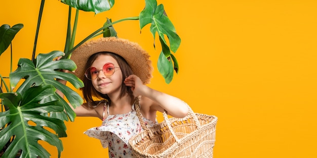 Menina bonitinha com óculos de sol laranja de chapéu de palha segurando uma bolsa de palha enquanto fica em pé em plantas tropicais