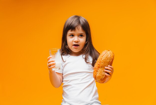 Menina bonitinha com um copo de leite e um pão em uma criança de fundo amarelo bebe comida saudável com leite