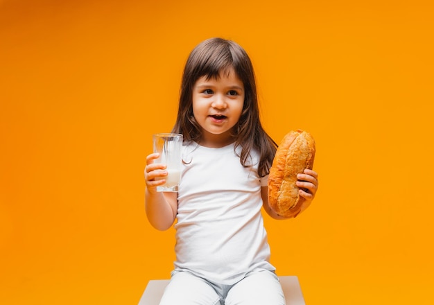 Menina bonitinha com um copo de leite e um pão em uma criança de fundo amarelo bebe comida saudável com leite