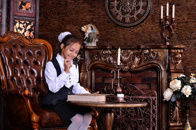 Menina bonitinha com uniforme escolar lendo livro na sala com móveis antigos