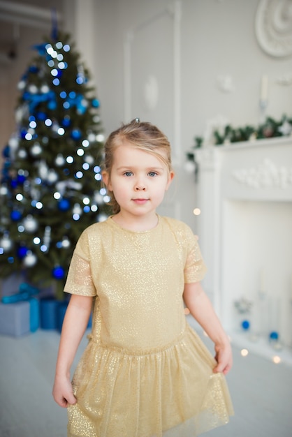 menina bonitinha com vestido dourado posando para o fundo da árvore de Natal, olhar para a câmera