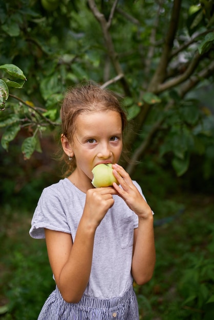 Menina bonitinha come maçã verde no jardim de casa, criança feliz ao ar livre